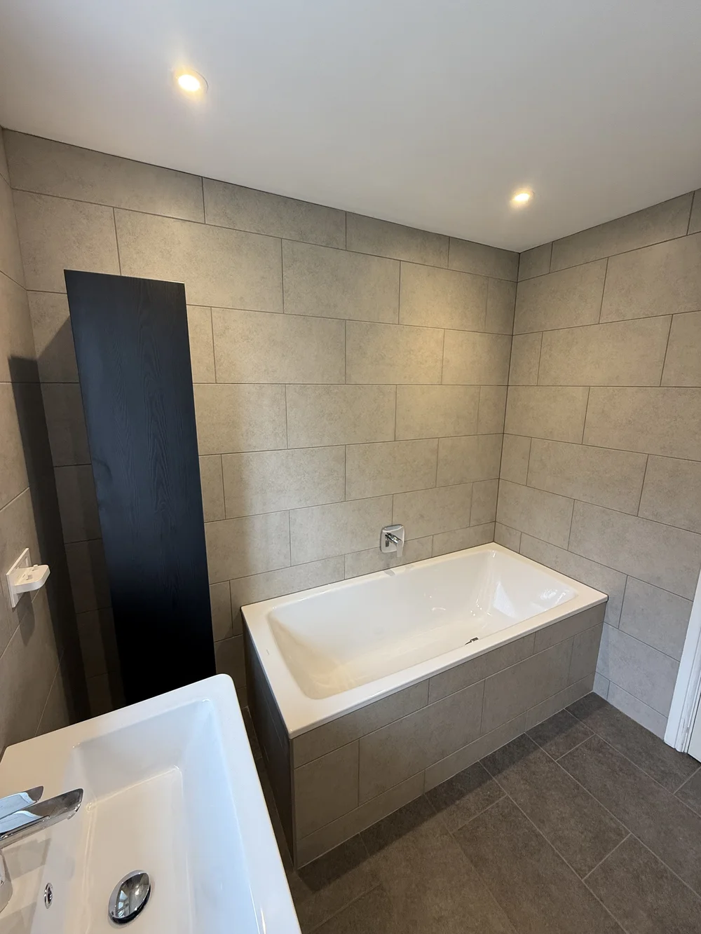 Stone-tiled bathroom with panelled bath and tall black storage column — bathroom renovation Haslemere