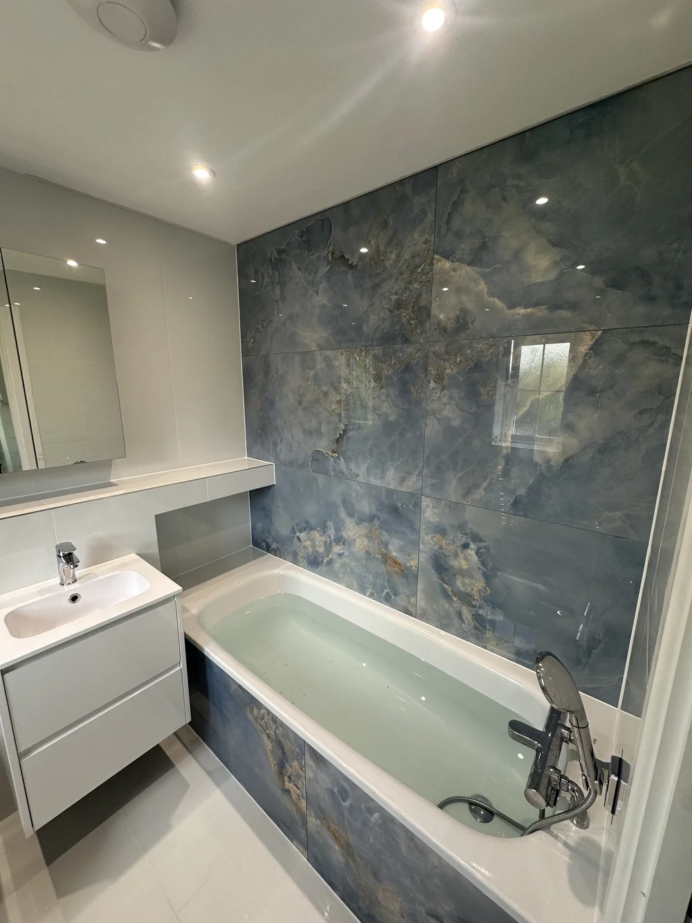 Family bathroom with blue onyx-effect feature wall, panelled bath and floating white vanity — bathroom installation Haslemere